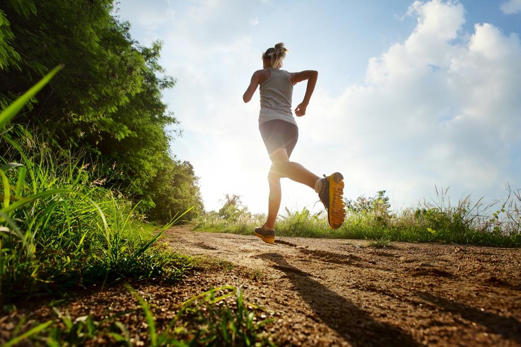 Osoba ubrana w sportowy strój biegnie po naturalnej, gruntowej ścieżce otoczonej zielenią. Ujęcie wykonane od tyłu, pod niskim kątem, podkreśla dynamikę ruchu. W tle widać błękitne niebo z rozproszonymi chmurami, a słońce świeci zza sylwetki biegacza, tworząc efektowną poświatę. Ruch