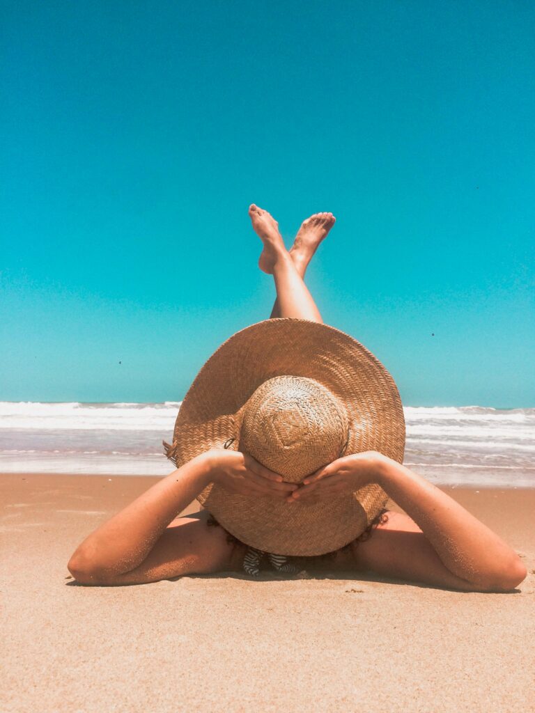 A woman in a straw hat relaxes on a sunny beach, enjoying the clear blue sky and ocean waves. lato