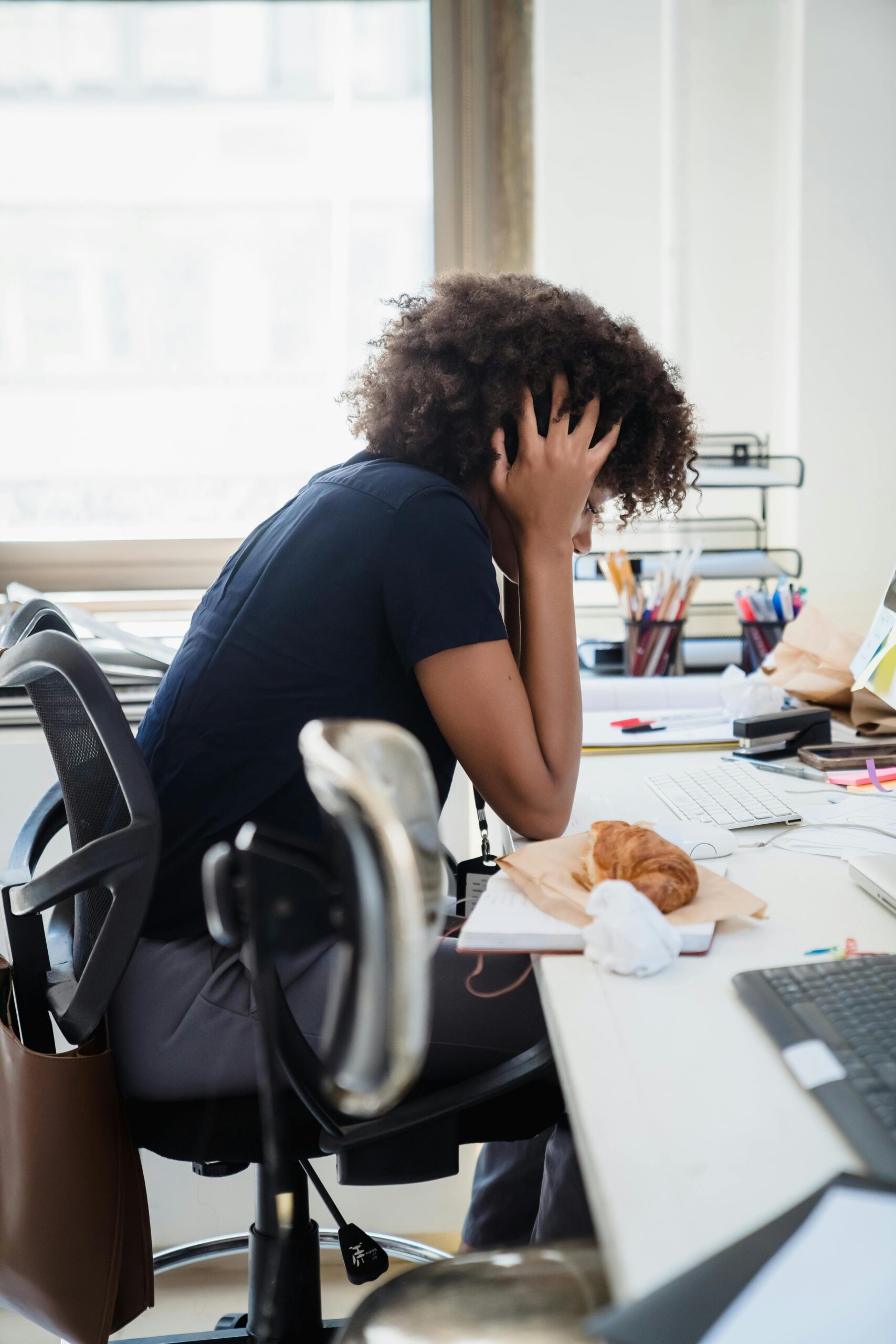 mgła mózgowa, A woman in an office holds her head in stress, croissant on desk.