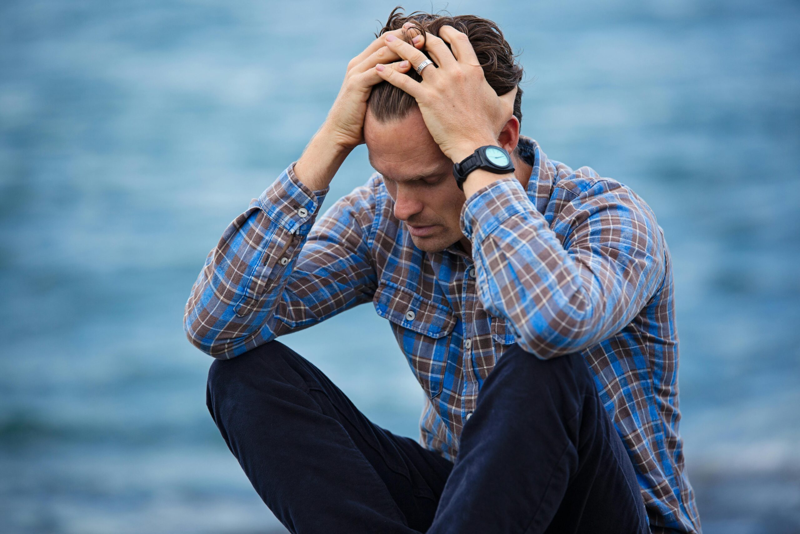 Zespół Cichego Przegrzania A man in a plaid shirt sits by the water looking distressed, symbolizing stress.